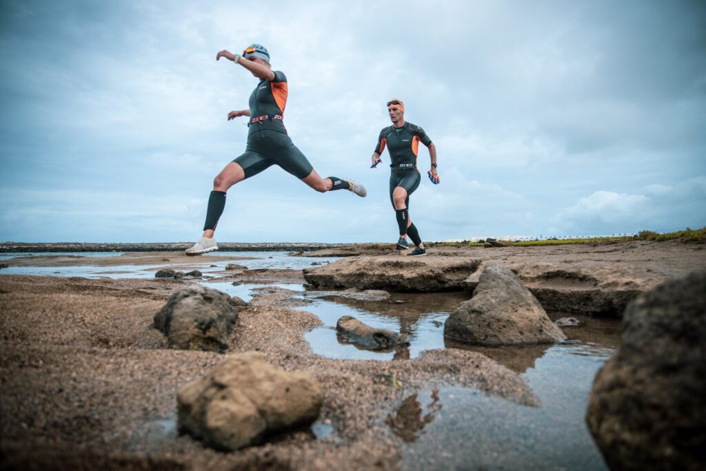 2 competitors wearing swimrun wetsuits jumping across rocks